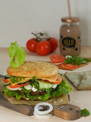 Burger with sesame bun, ham and vegetables, сucumber, tomato, cheese, onion, sauce and salad, lies on a dark cutting board on a white background. Latte in a jar with a straw. Tomatoes in the back.