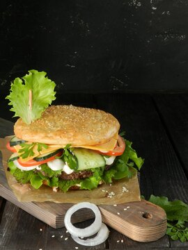 sandwich with ham and vegetables. Burger with sesame bun, cucumber, tomato, cheese, onion, sauce and salad, lies on a dark cutting board on a black background. Photo for the recipe. Photo for the menu