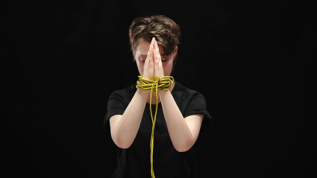 Clasped female hands tied with rope with blurred sad woman at background. Young Caucasian victim of human trafficking posing at black background. Humiliation and tyranny concept.