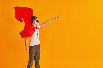 cheerful child is playing in a superhero costume. the boy is dressed in a red cloak and mask, holding out his hand on a yellow background. the concept of a superpowered boy striving for victory