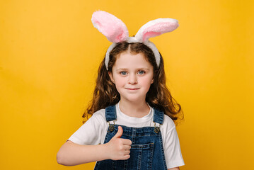 Close up head shot portrait of smiling little girl in pink bunny fluffy ears. Happy kid liking and thumb up on yellow studio background, child looking at camera, giving recommendation. Easter concept