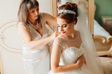 The mother helps the slender bride to lace up her white wedding dress, buttoning up in a delicate lace pattern with a fluffy skirt at the waist. Morning wedding preparation details of the newlyweds