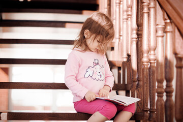 Studious little girl sitting on wooden stairs and reading book