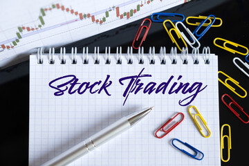 On the desktop are a forex chart, paper clips, a pen and a notebook in which it is written - Stock Trading