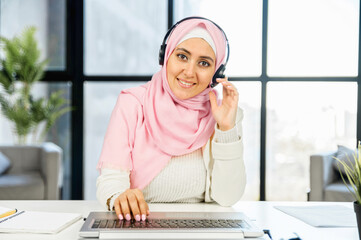 Smiling arabian woman operator in hijab, customer support call in office, sitting with headset, typing on the keyboard touching the earpiece with the hand, there's no query she can't solve, love job