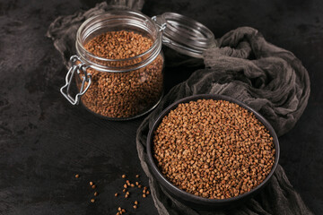 Organic uncooked scattered buckwheat grain in a bowl and glass jar on a black background