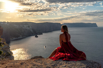Fototapeta na wymiar A girl with loose hair in a red dress sits on a rock rock above the sea. In the background, the sun rises from behind the mountains.