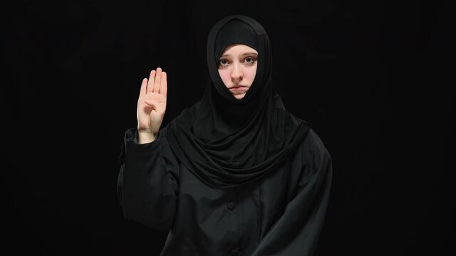 Middle shot of depressed terrified Muslim woman raising palm with tuck thumb looking at camera. Portrait of oppressed victim of domestic violence posing at black background showing sign to help.