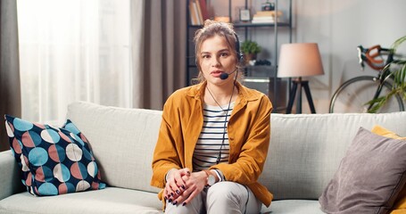 Obraz premium Beautiful young happy Caucasian female in headset speaking looking at camera while sitting on sofa in living room at home. Woman freelancer working talking on online web conference, e-learning concept