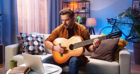 Joyful young Caucasian bearded man having online video lesson on laptop computer learning how to play the guitar while sitting on couch in living room, musician playing the instrument at home