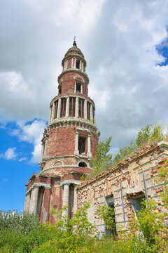 an abandoned brick bell tower with columns, a cross and part of a destroyed wall overgrown with greenery, in the background, against the sky, grass in the foreground