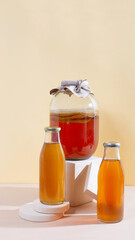 Glass gar with komucha tea and two bottles with ready beverage on modern podiums in front of yellow background with copy space. Healthy fermented drink. Vertical