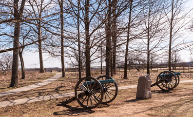Fototapeta na wymiar Cannons and a stone monument in a wooded grove with a walkway through it in the Gettysburg National Military Park on a sunny spring day