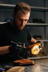 Portrait of bearded shoemaker wearing black gloves spraying paint of light brown leather shoes, close-up. Concept of cobbler artisan repairing and restoration work in shoe repair shop.