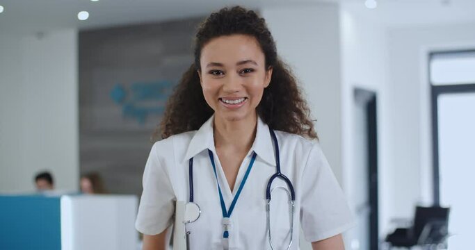 Portrait satisfied african american female doctor in white medical coat looking at camera and smiles standing in hospital. Cheerful young mixed race woman, curly black hair nurse