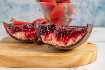 Two sliced ​​stuffed easter egg pieces, with a juicy filling of strawberry jam layer, biscuit layer, chocolate and cream. And a bowl full of strawberries in the background