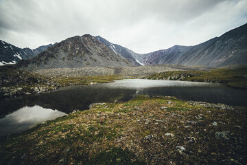 Scenic mountain landscape with glacial lake among mountains under gray cloudy sky. Atmospheric alpine scenery with transparent mountain lake in rainy weather. Beautiful clear alpine lake among rocks.