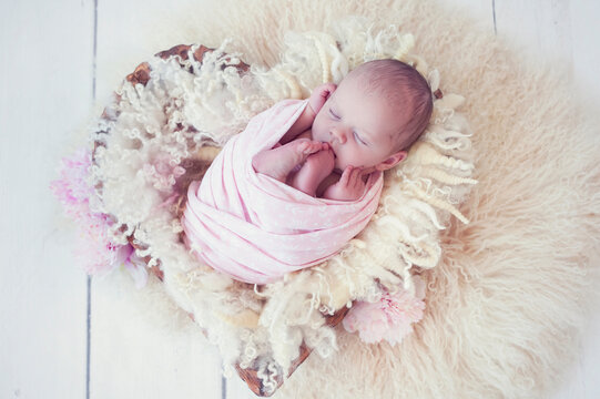 Newborn baby girl wrapped in pink wrap on wool fluffy blanket in wooden bowl in the shape of heart on white wood floor. Sweet infant sleeping in props for newborn photography