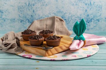 Four small chocolate-flavored brigadier cakes on an Easter table with pink napkin, napkin ring similar to rabbit ears, a themed sousplat and wooden stand. Blue background