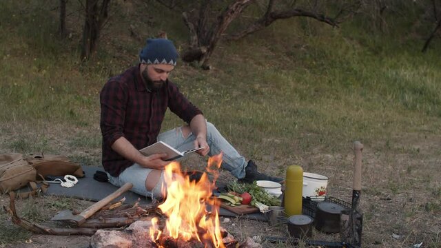 Traveler man sitting near campfire and reading book. Hiker tourist resting near bonfire at camp, camping in woods in evening. Male camper relaxes on vacation in wild nature