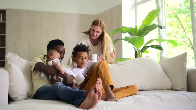 Mixed biracial family use tablet together, mother stand and watch and dad with kids sit on sofa Spbd
