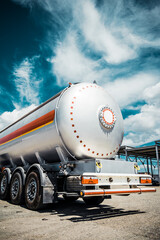 Truck with trailer, tank with flammable liquid, sunny day outside, metallic color container, blue sky with white clouds, gravel