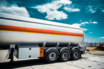Truck with trailer, tank with flammable liquid, sunny day outside, metallic color container, blue sky with white clouds, gravel