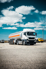 Fototapeta na wymiar Truck with trailer, tank with flammable liquid, sunny day outside, metallic color container, blue sky with white clouds, gravel