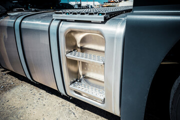 Truck with trailer, tank with flammable liquid, sunny day outside, metallic color container, blue sky with white clouds, gravel