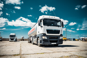 Truck with trailer, tank with flammable liquid, sunny day outside, metallic color container, blue sky with white clouds, gravel
