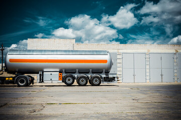Truck with trailer, tank with flammable liquid, sunny day outside, metallic color container, blue sky with white clouds, gravel