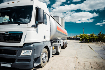 Truck with trailer, tank with flammable liquid, sunny day outside, metallic color container, blue sky with white clouds, gravel