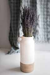 Bouquet of dry lavender flowers in a rustic clay vase over a white table with a hung, blurred, turquoise blanket at the background.