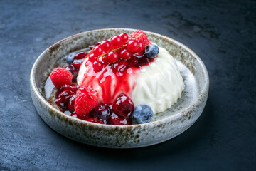Modern Style traditioneller Mandelsulz Pudding mit Wildbeeren Coulis serviert als close-up in einem Nordic Design Teller auf schwarzen Hintergrund