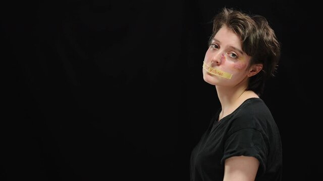 Abused beaten woman with bruises on face turning to camera with mouth closed with Stop violence sign. Portrait of oppressed Caucasian victim of domestic tyranny posing on the right at black background