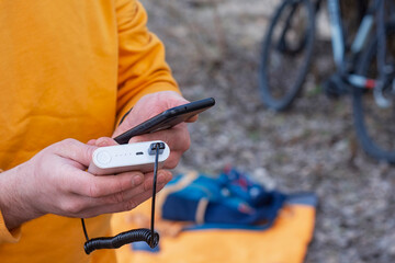A tourist charges a smartphone with a power bank on the background of a backpack and a bicycle in nature.