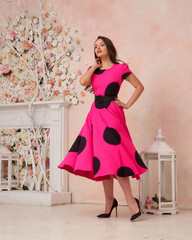 Pretty beautiful young woman with long brunette wavy hair wearing midi polka dot pink and black dress posing in bright interior