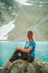 A beautiful young woman in trekking gear sits on a rock near a turquoise lake in the mountains. Mountain peaks, snowfields, and a glacier are in the background. Portrait from the back