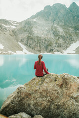 A beautiful young woman in trekking gear sits on a rock near a turquoise lake in the mountains. Mountain peaks, snowfields, and a glacier are in the background. Portrait from the back