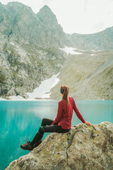 A beautiful young woman in trekking gear sits on a rock near a turquoise lake in the mountains. Mountain peaks, snowfields, and a glacier are in the background. Portrait from the back