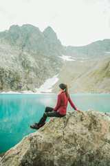 A beautiful young woman in trekking gear sits on a rock near a turquoise lake in the mountains. Mountain peaks, snowfields, and a glacier are in the background. Portrait from the back