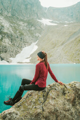 A beautiful young woman in trekking gear sits on a rock near a turquoise lake in the mountains. Mountain peaks, snowfields, and a glacier are in the background. Portrait from the back