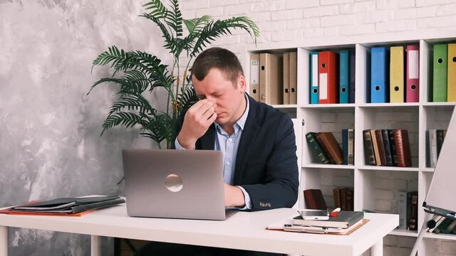 Eye fatigue, poor eyesight, office work. A young man takes off his glasses and rubs his eyes with his hand while sitting at a table with a laptop in a suit.