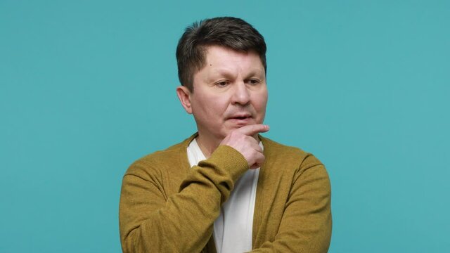 Hmm, need to think. Doubtful bewildered middle aged man in white t-shirt and cardigan contemplating rubbing his chin, hesitating making choice. Indoor studio shot isolated on blue background