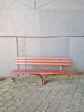 Empty Brown Wooden Bench On Footpath Against Grey Brick Wall With Tiled Floor.