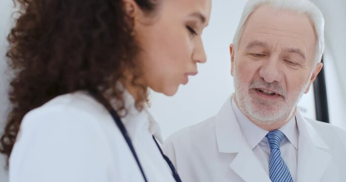 Portrait two medical workers aged caucasian male and young curly hair multiethnic female, doctors discussing medical case, talking about health report while standing by white wall in hospital