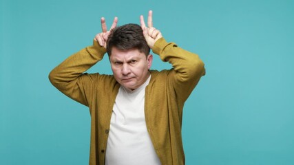 Dangerous arrogant middle aged dark haired man holding hands above head showing bull horns, looking at camera with aggression, ready to attack. Indoor studio shot isolated on blue background - Powered by Adobe
