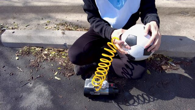 Close up image showing a caucasian woman holding a soccer ball and inserting needle bit at the end of the yellow coiled tubing attached to 12v car tire inflator to pump in air to the ball. 