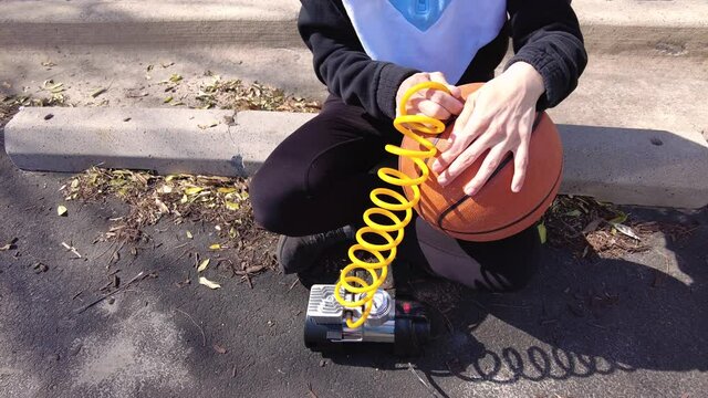 Close up image showing a caucasian woman holding a basketball and inserting needle bit at the end of the tubing attached to 12v car tire inflator to pump in air to the ball. 