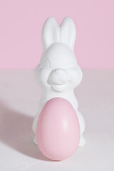 White Easter bunny with a pink Easter egg on a concrete table, on a pink background. Hard shadows. Vertical orientation. Copy space